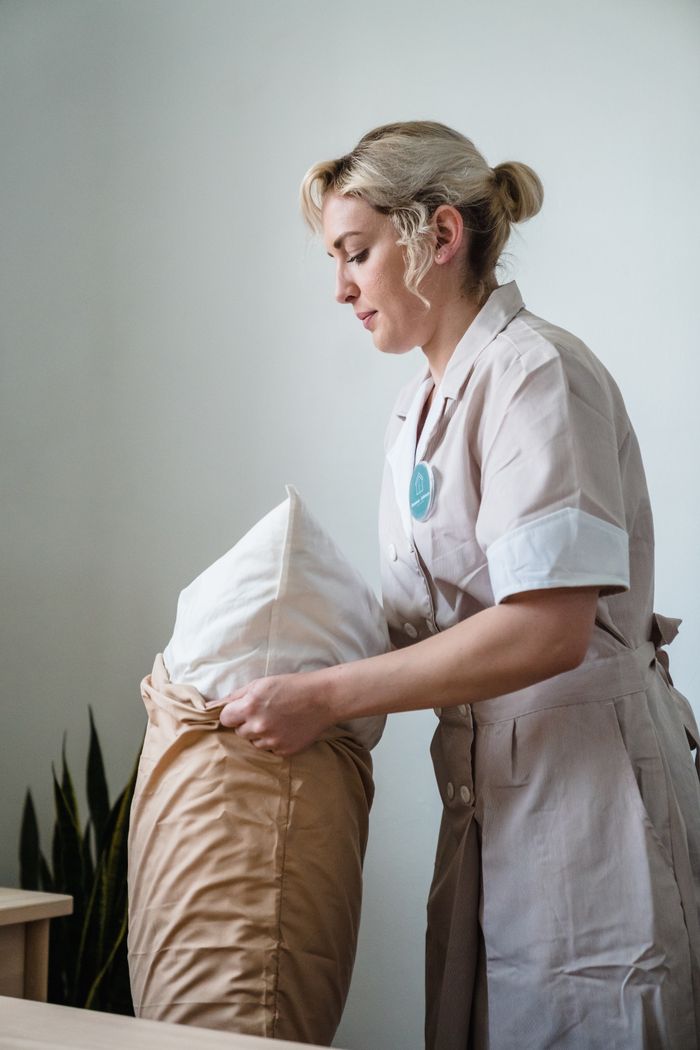 gallery-3 Hotel maid in uniform arranging pillows in a neatly styled guest room.