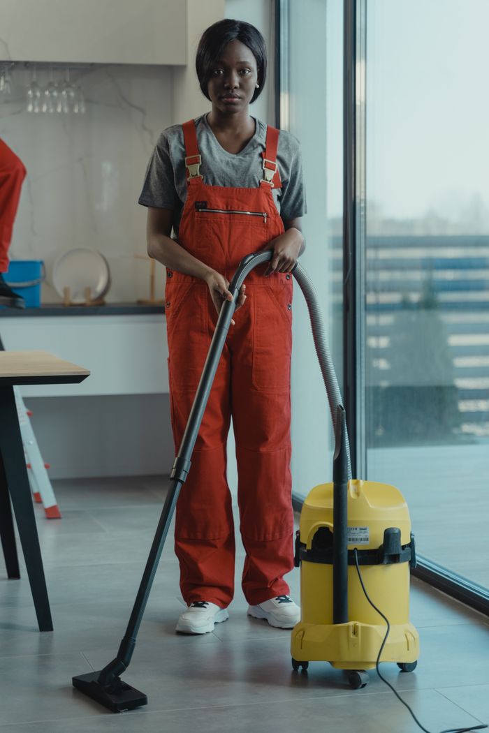 hero-img-01 An adult woman in red coveralls is using a vacuum cleaner in a modern indoor space, with attention on cleaning duties.